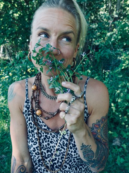 Karoline Karttunen från Karolines way of Life håller blommor av jordreva framför ansiktet. Hon tittar åt sidan och ler. Grönt