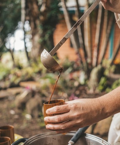 Man häller upp ceremoniell kakao i kopp av trä för kakaoceremoni. Han häller upp kakaodrycken från en stor kastrull under koppen och det står fler koppar vid sidan av.