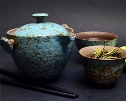 En blå turkos tekanna i keramik med lock står på mörk bakgrund. Bredvid står två tekoppar med olika mönster på utsidan och brunt på insidan. I koppen längst fram ligger det örter. På bordet ligger ockås två pinnar, ätpinnar.